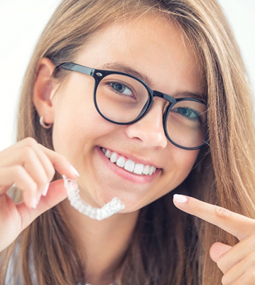 A young girl with glasses pointing to her smile and the aligner she’s holding in her hand as part of her Invisalign in Richardson treatment