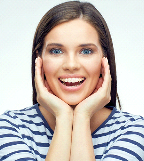 A young woman wearing a blue and white striped shirt holds her face between her hands while showing off her clear braces