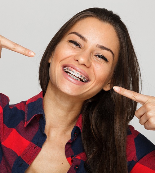 A woman wearing a plaid shirt and pointing to her smile that is being treated with metal braces by her orthodontist in Richardson