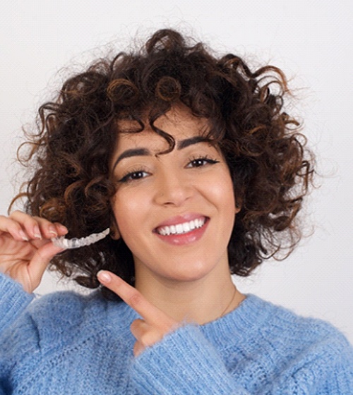 A young woman wearing a blue sweater and holding a clear aligner after learning about the cost of Invisalign in Richardson