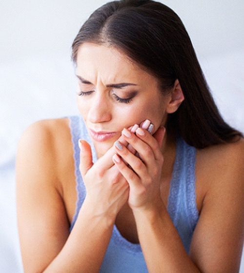 A young woman sitting on her bed and holding her cheek in pain because of her braces