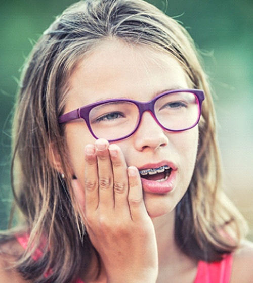 A young girl wearing glasses and braces holding her mouth in pain
