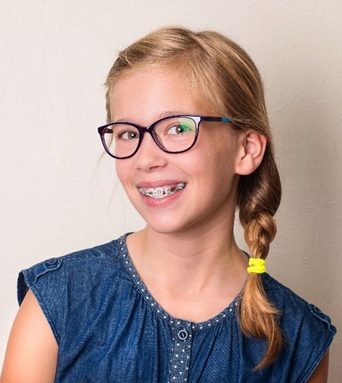 A young girl wearing a blue blouse and glasses is smiling and showing off her metal braces