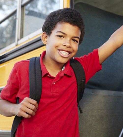 A school-aged boy wearing a red polo shirt and backpack stands at the door of a bus and smiles while wearing his braces